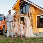 Family of four happily standing outside their modern wooden house on a sunny day.